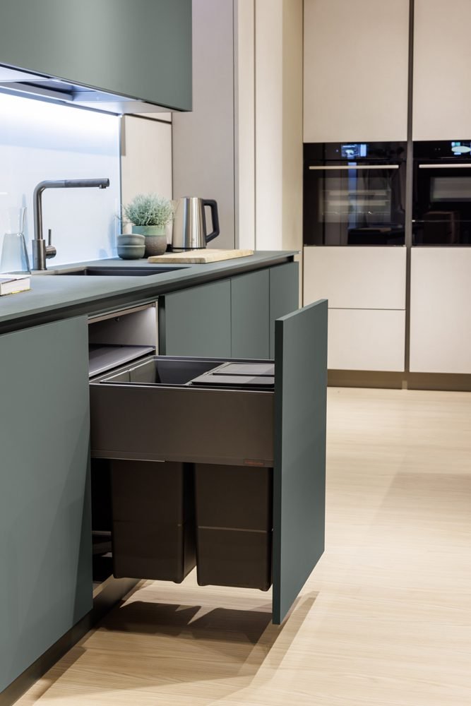 A sleek, modern kitchen featuring matte sage green cabinetry with a pull-out waste management system. The open drawer reveals two integrated dark grey trash bins. In the background, there is a minimalist sink with a matte grey faucet, a stainless steel kettle, and built-in black double ovens set into light beige wall units.