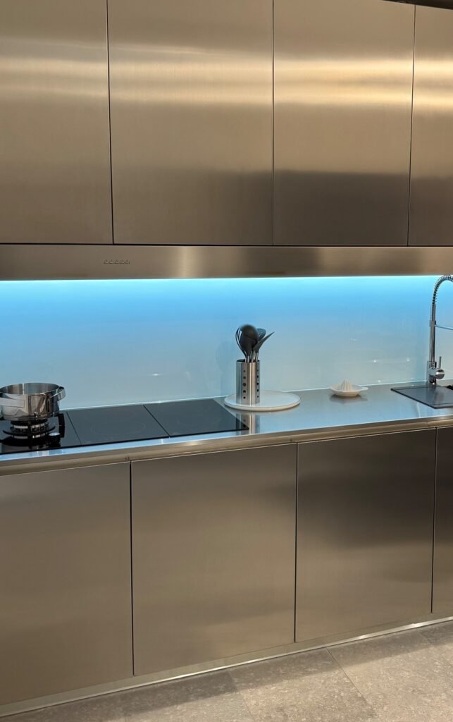 Modern stainless steel kitchen cabinets with brushed metal finish, sleek countertop, integrated cooktop, and illuminated glass backsplash.
