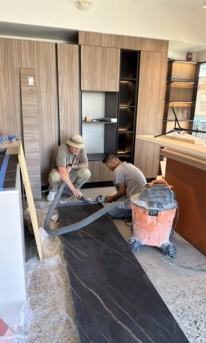 Craftsmen installing custom Italian cabinetry and a dark stone countertop in a modern European kitchen showroom with wood finishes and built-in shelving