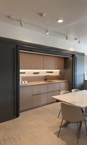 Modern Italian kitchen showroom display with light wood cabinetry, integrated lighting, dark sliding panels, a sleek sink area, and a marble-style dining table in the foreground.