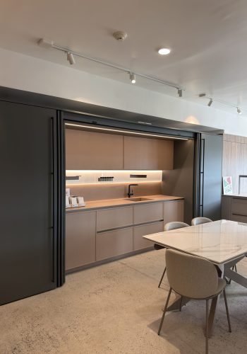 Modern Italian kitchen showroom display with light wood cabinetry, integrated lighting, dark sliding panels, a sleek sink area, and a marble-style dining table in the foreground.