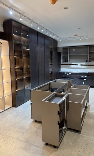 Italian cabinetry showroom with custom storage displays, tall dark wood cabinets, open shelving, track lighting, and partially installed cabinet modules.