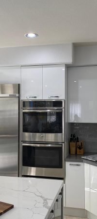 White kitchen with stainless steel refrigerator, built-in ovens, range hood, white cabinetry, stone countertops, and a central island.