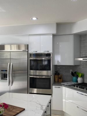 White kitchen with stainless steel refrigerator, built-in ovens, range hood, white cabinetry, stone countertops, and a central island.