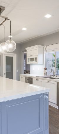 White kitchen with blue island, pendant lighting, glass cabinet doors, wood flooring, and a bright traditional coastal design.