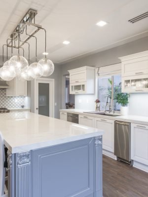 White kitchen with blue island, pendant lighting, glass cabinet doors, wood flooring, and a bright traditional coastal design.